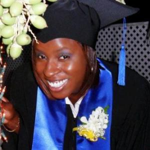 A graduate wearing a black cap and gown, smiling brightly, with a blue sash and a white flower corsage.
