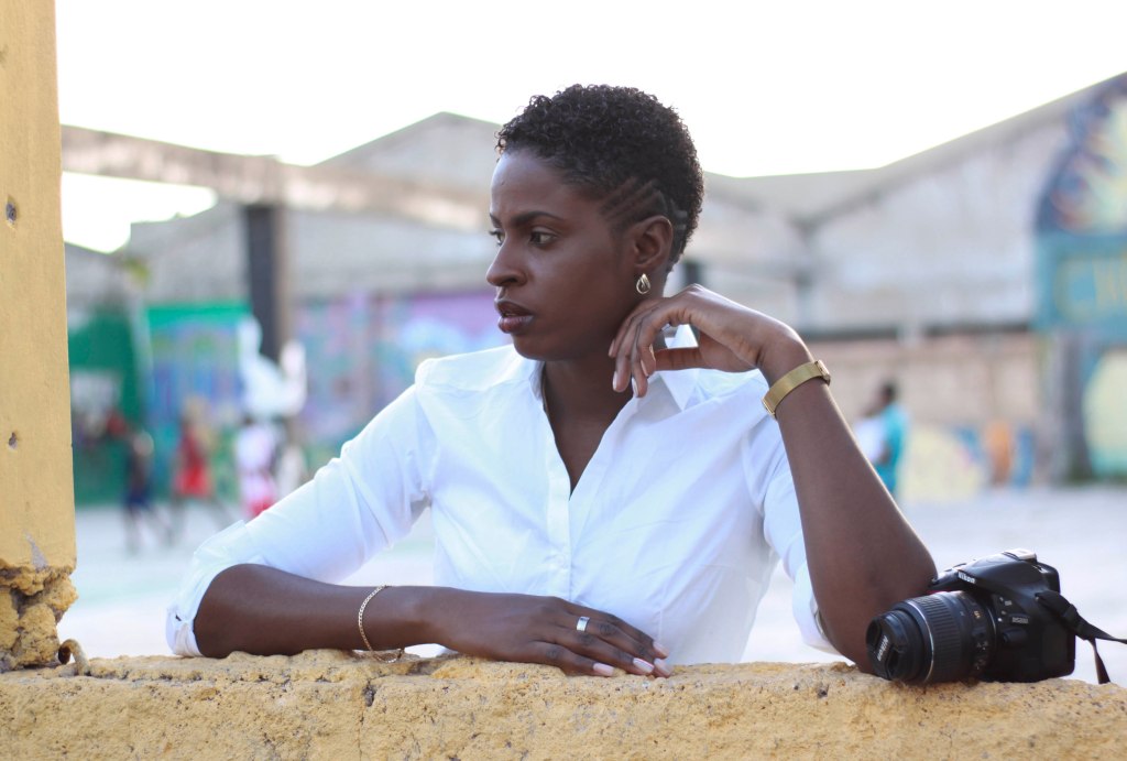 A woman in a white shirt leans against a yellow wall, looking thoughtfully off to the side, with a camera resting nearby.