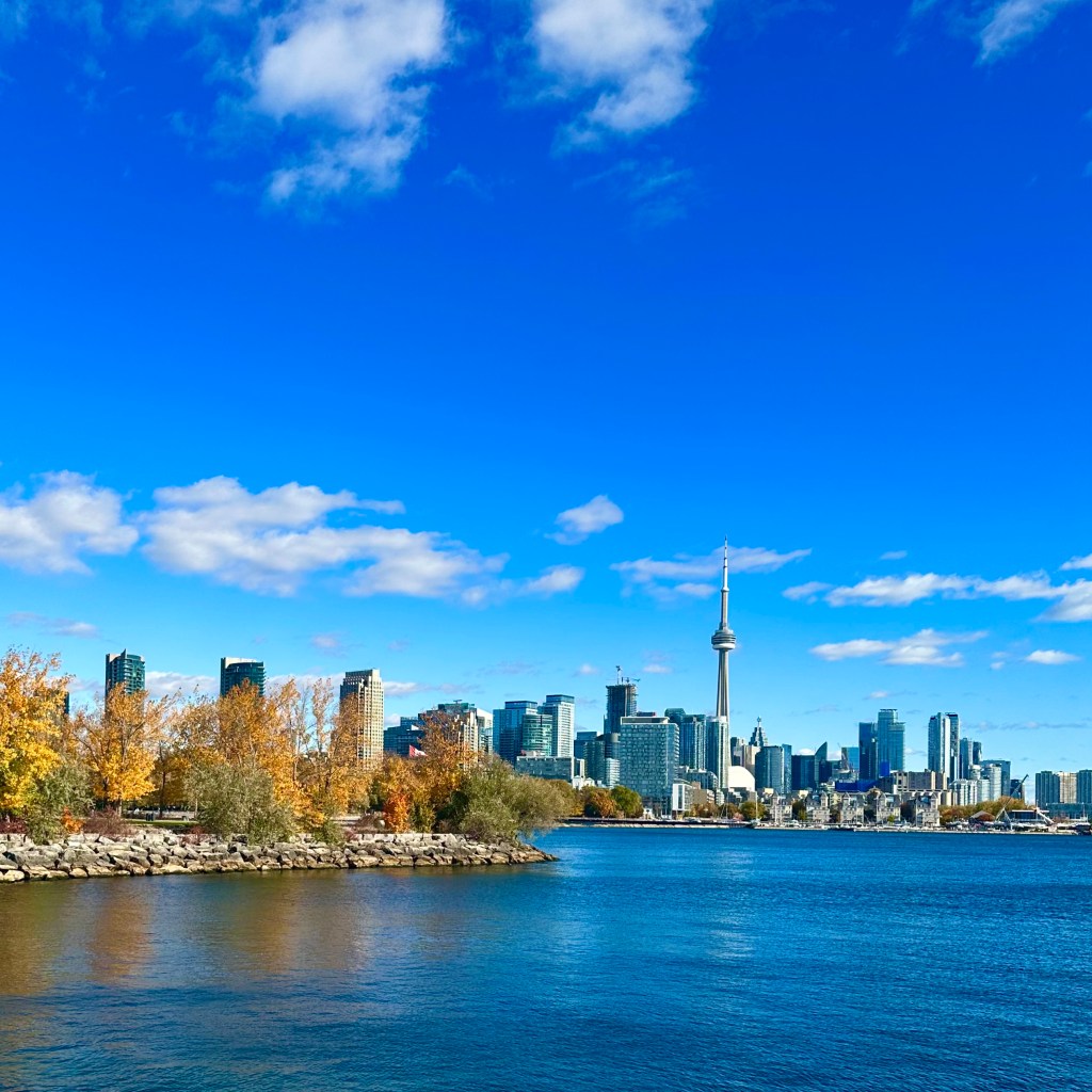 A panoramic view of a city skyline featuring the CN Tower and colorful autumn foliage along the waterfront, under a bright blue sky with fluffy white clouds.