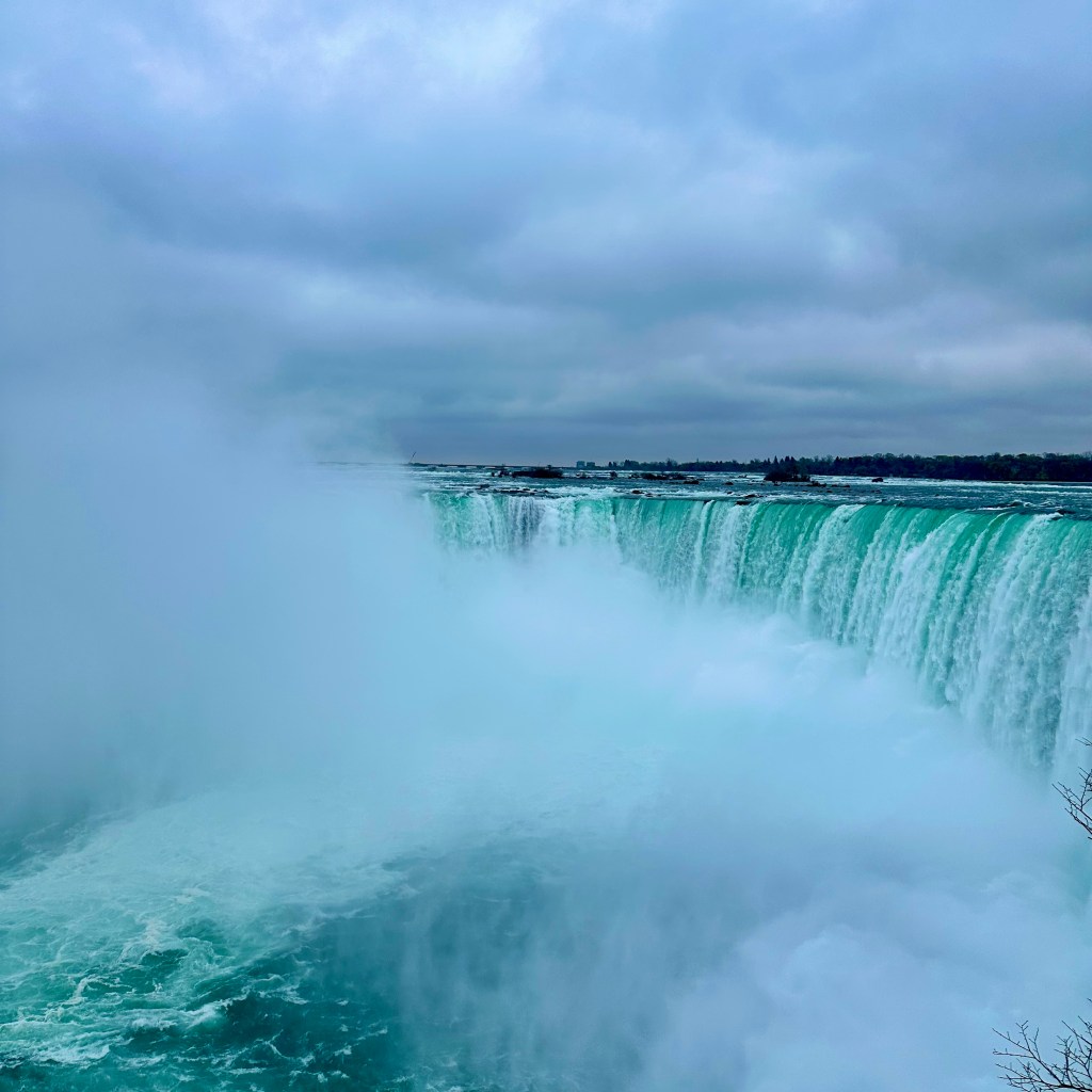 A dramatic view of Niagara Falls, showcasing the cascading water plunging into a misty pool below, under a cloudy sky.