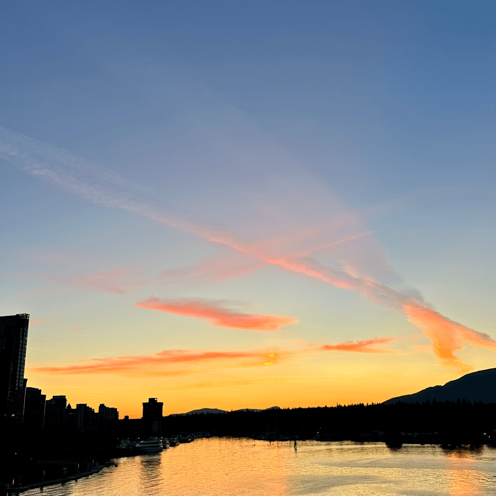 A panoramic view of a sunset with pink and orange clouds above calm waters, flanked by silhouetted buildings on one side and mountains on the other.
