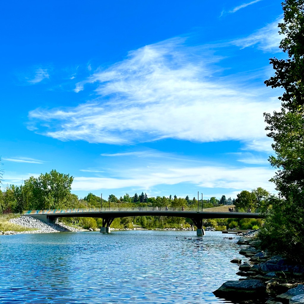 A scenic view of a bridge spanning over a calm river, surrounded by lush greenery and a clear blue sky with wispy clouds.