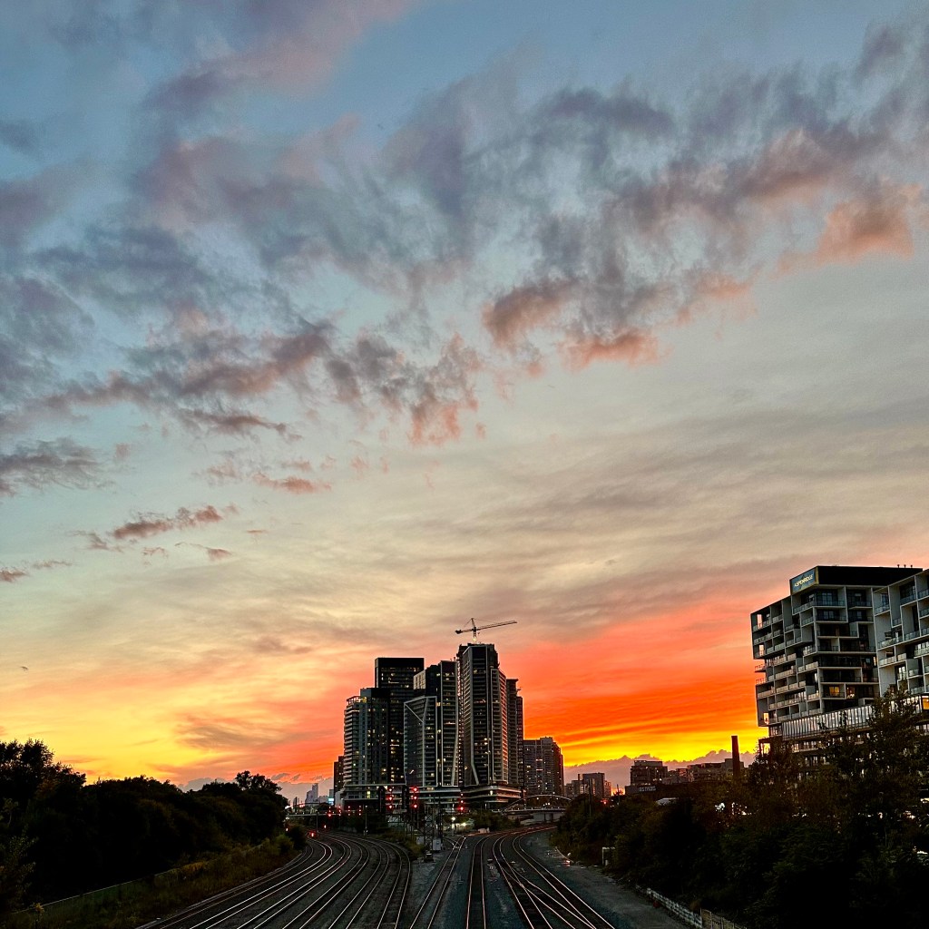 A panoramic view of a city skyline at sunset, featuring buildings silhouetted against vibrant orange and blue skies with scattered clouds, and train tracks in the foreground.