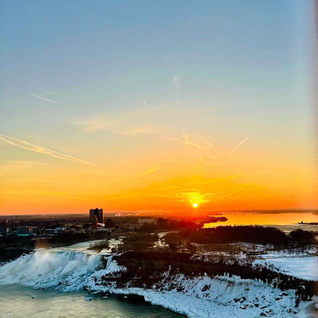 A scenic view of a sunset over a frozen waterfall, with a vibrant orange sky and silhouettes of buildings and trees in the foreground.