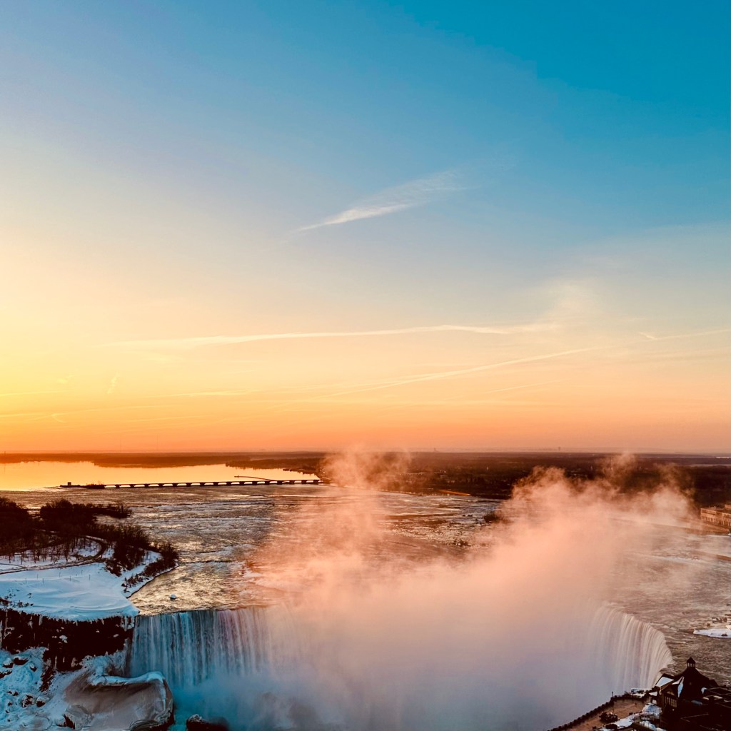 A panoramic view of Niagara Falls at sunset, with vibrant orange and blue skies, mist rising from the falls, and a distant bridge over a calm waters.