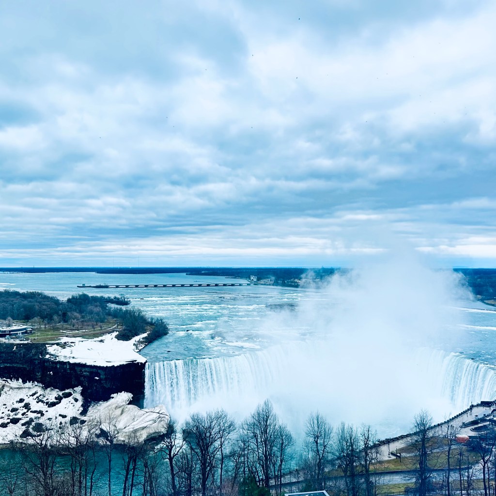 A view of Niagara Falls with icy edges and mist rising, under a cloudy sky.