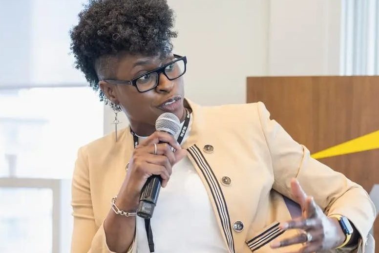 An executive leader speaking into a microphone during a panel discussion, wearing glasses and a beige jacket, with a background featuring a modern office setting.